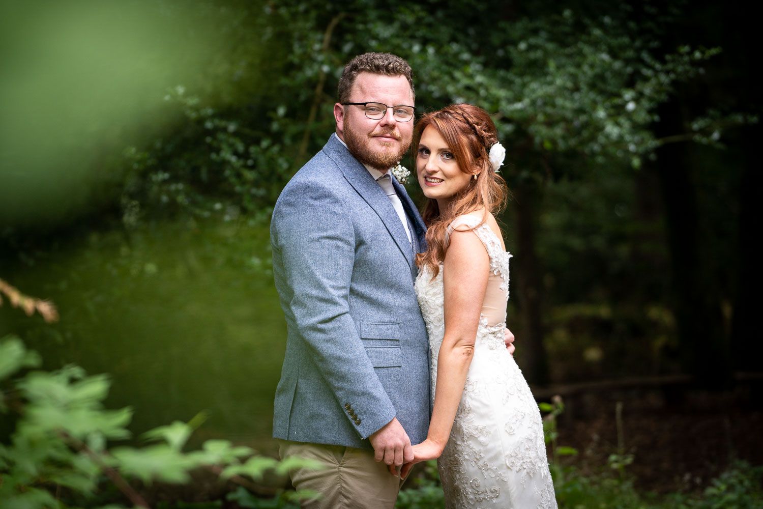 A couple poses together outdoors in wedding attire with a blurred green nature backdrop.