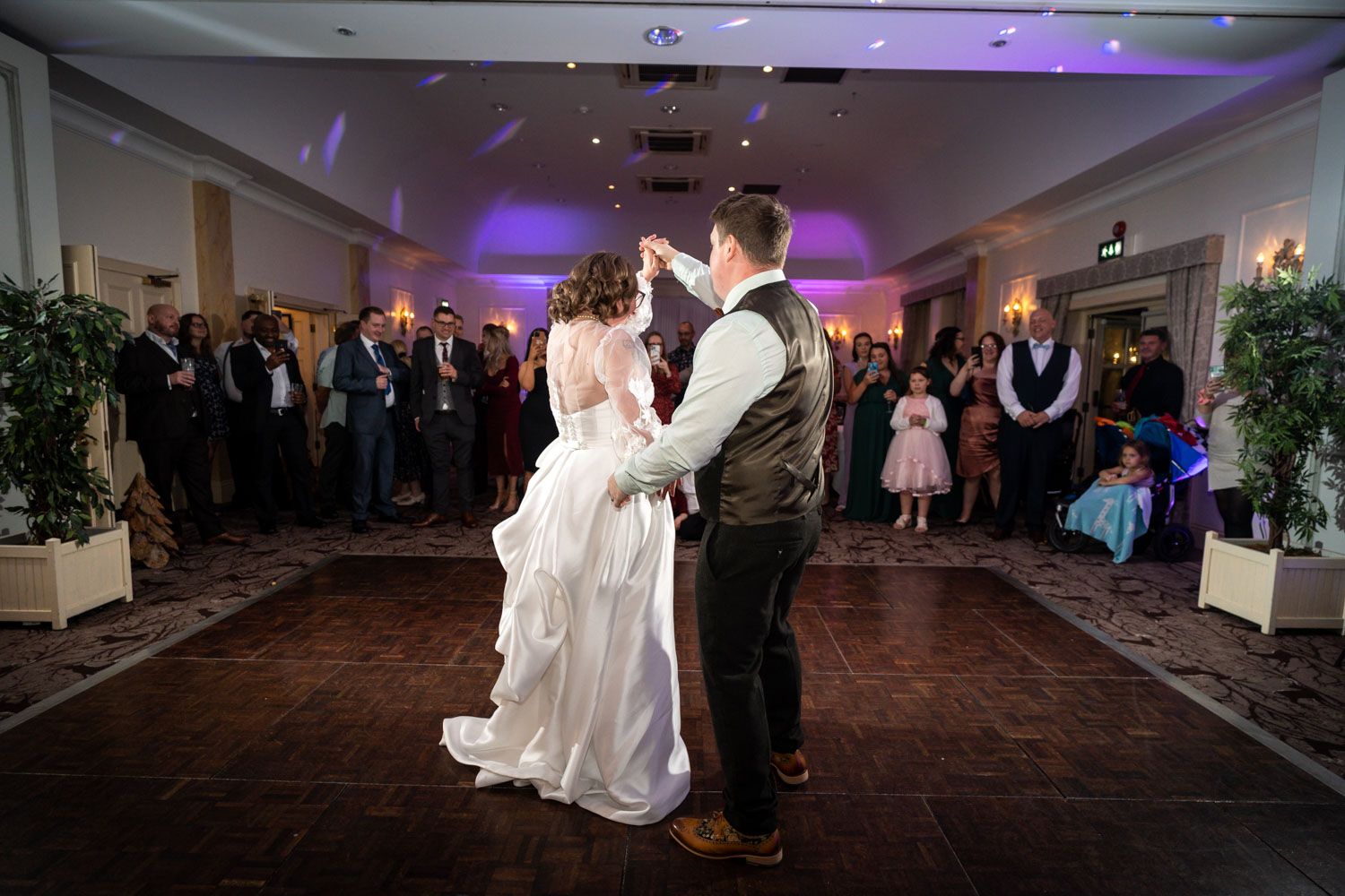 A couple shares their first dance as newlyweds on a dark wooden dance floor surrounded by wedding guests.