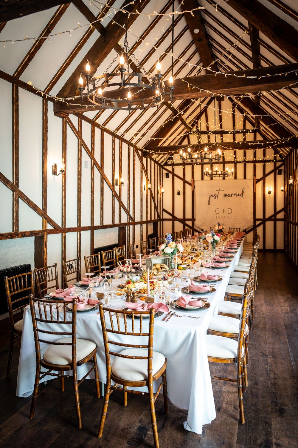 Long rustic barn wedding reception table with white linens and wooden chairs beneath exposed wooden beams and chandelier.