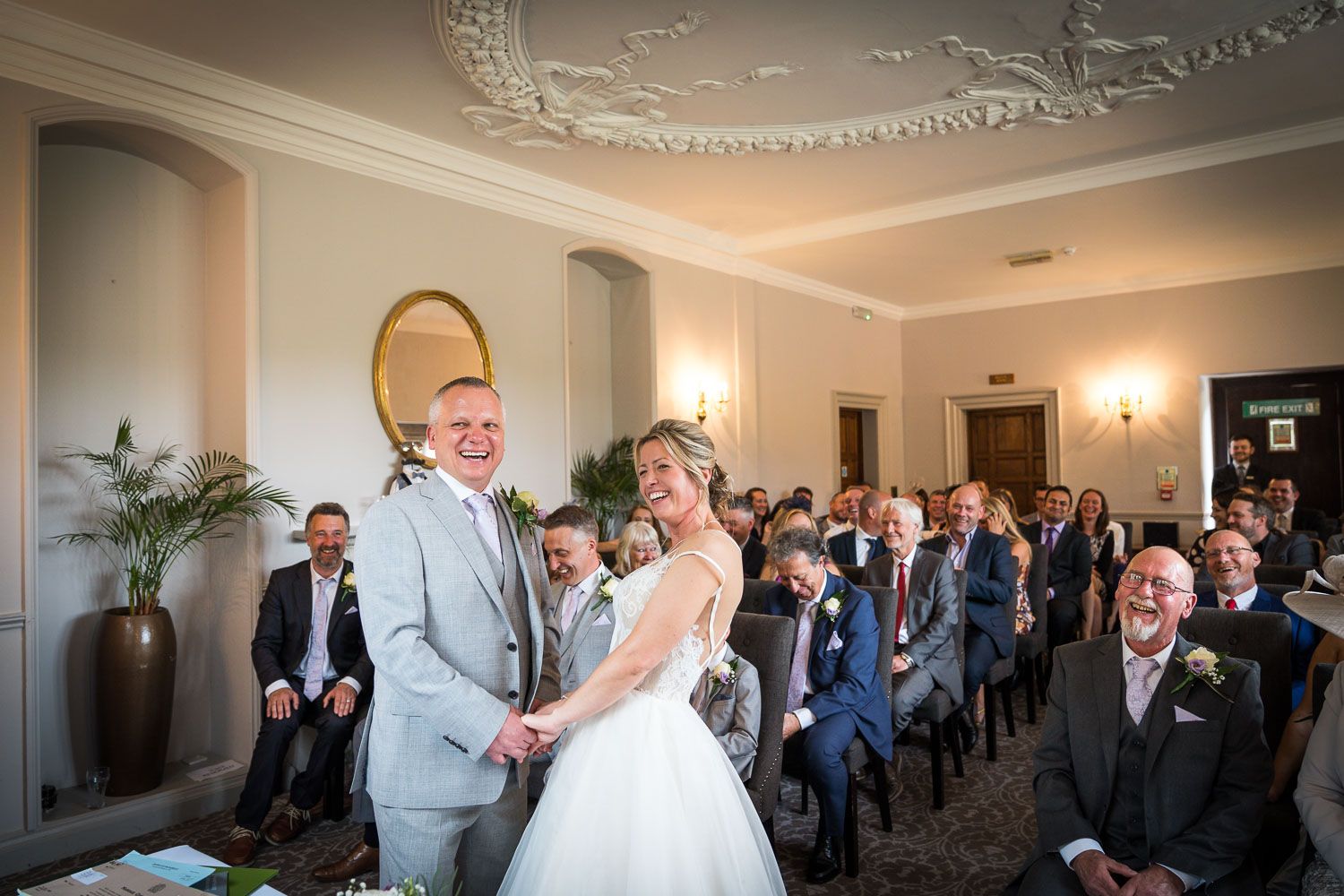 Wedding ceremony in elegant room with ornate ceiling as couple shares joyful moment at the altar.