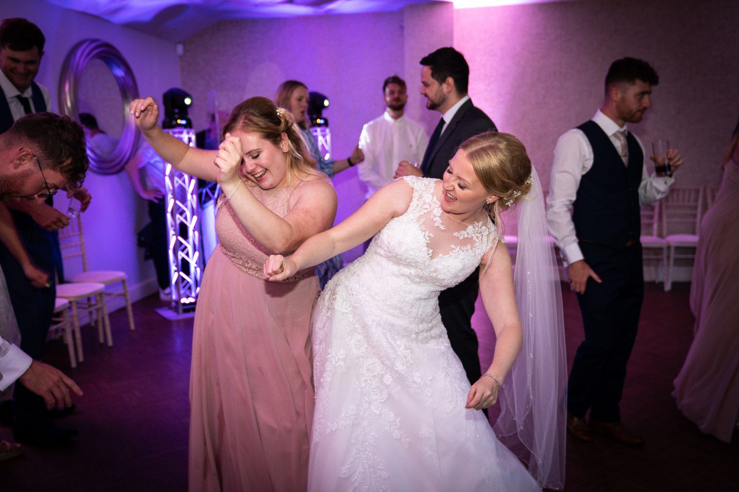 Dancing guests celebrate at a wedding reception with pink and purple lighting in an elegant venue.