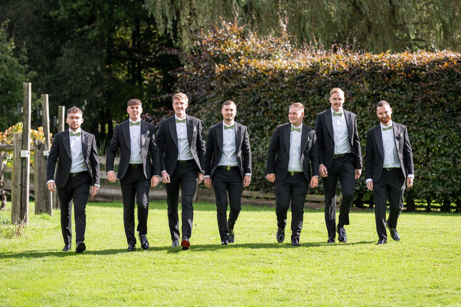 Seven groomsmen in matching black tuxedos and bowties walk in sync across a sunny green lawn at an outdoor venue.