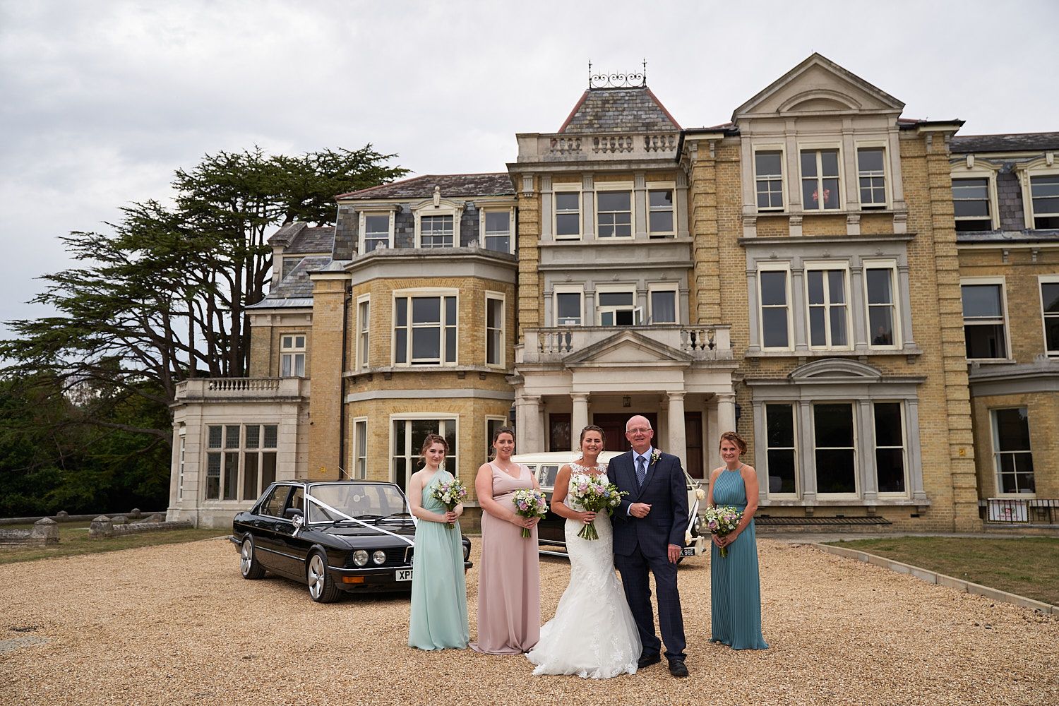 Wedding party poses in front of an elegant Victorian mansion with a classic car parked nearby.