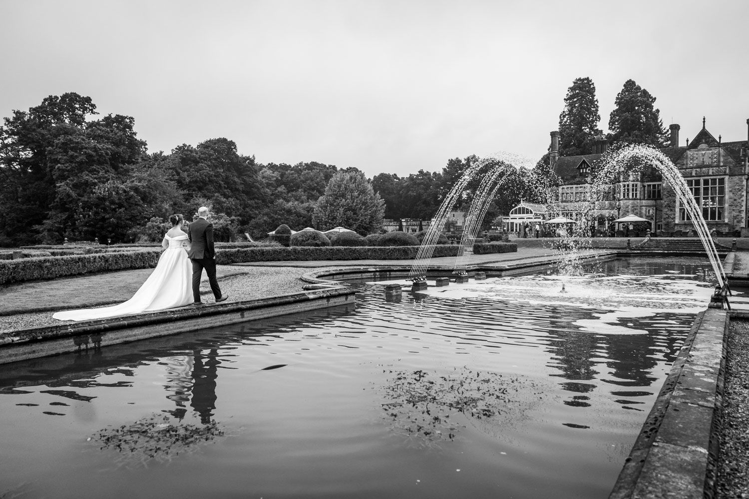 Elegant black and white photo of newlyweds by a reflecting pool with arching water fountains at a formal garden.