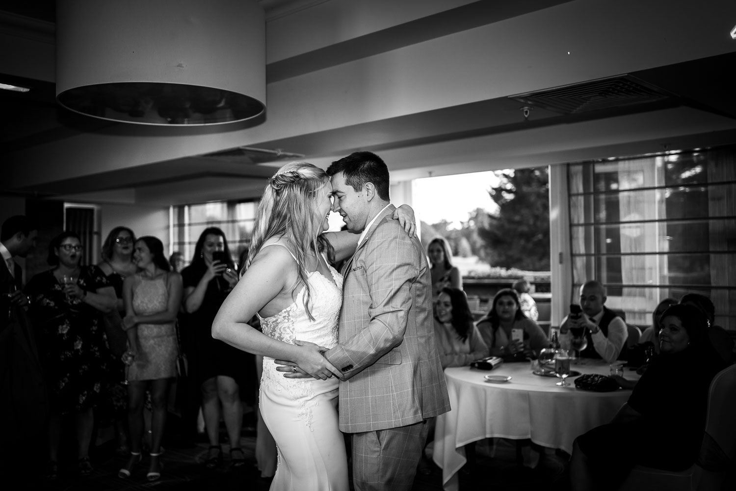 A wedding couple shares their first dance together in black and white while guests watch from the reception venue.