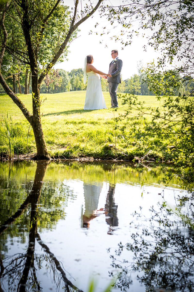 Wedding reflection in still pond water with couple standing in sunlit grassy field framed by trees on summer day.