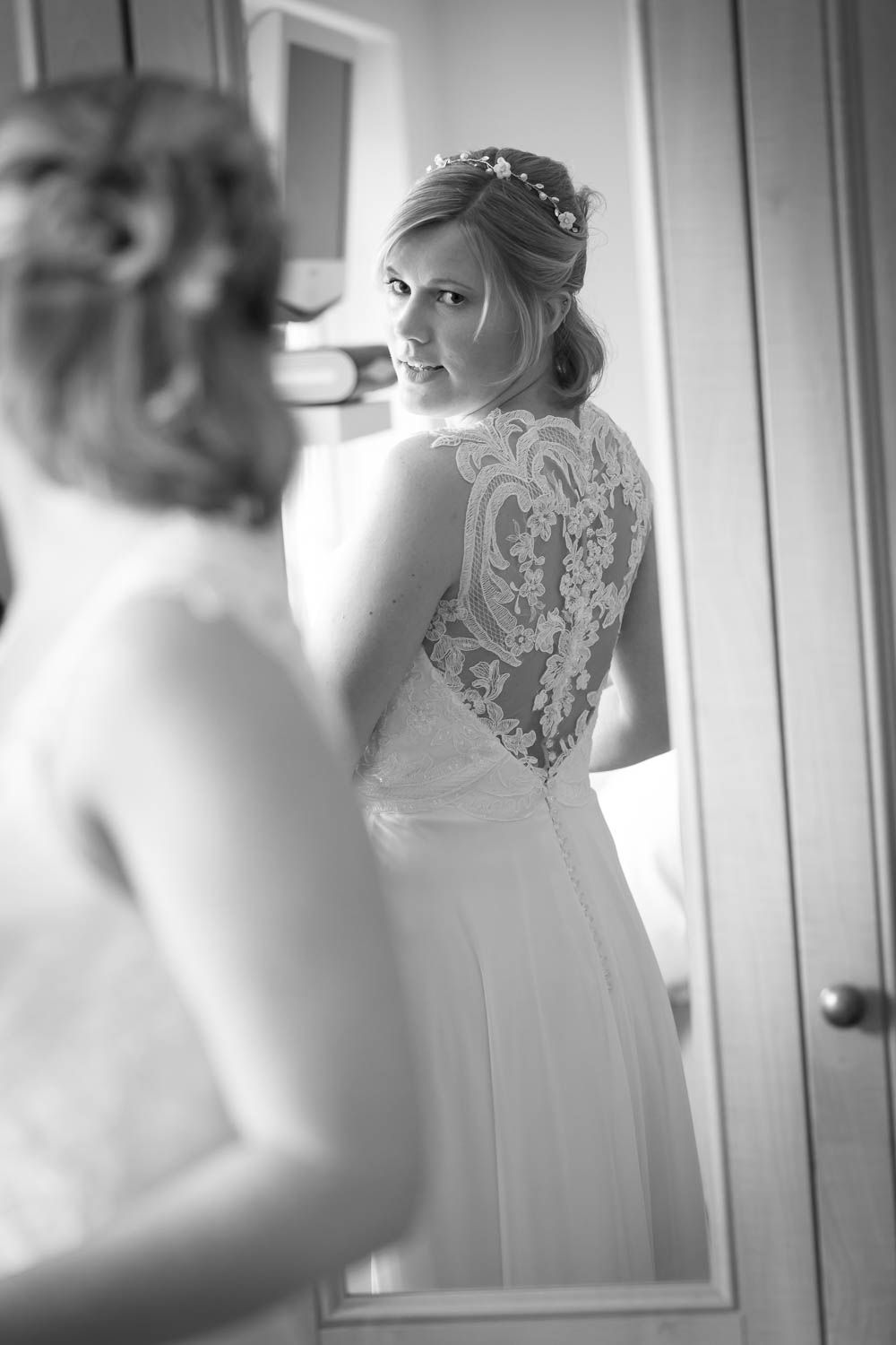 A bride in a flowing white gown with delicate lace detailing on the back looks over her shoulder in a black and white photo.