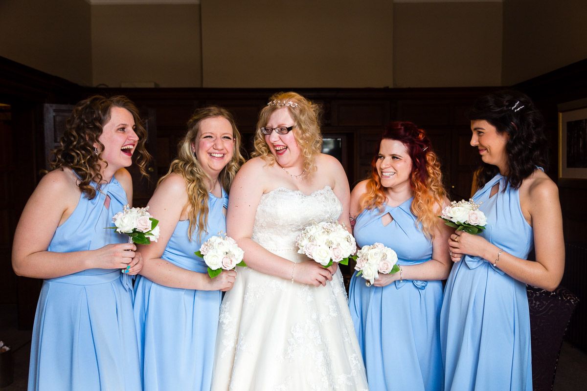 Bridesmaids in light blue dresses share a laugh with bride in white gown while holding white bouquets.