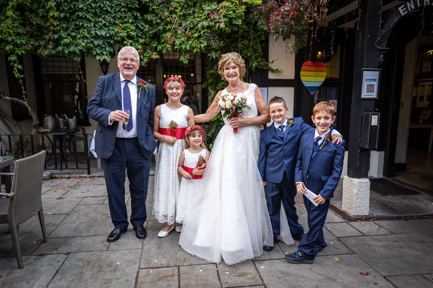 A group in formal wedding attire smile together outside a brick building with climbing vines.