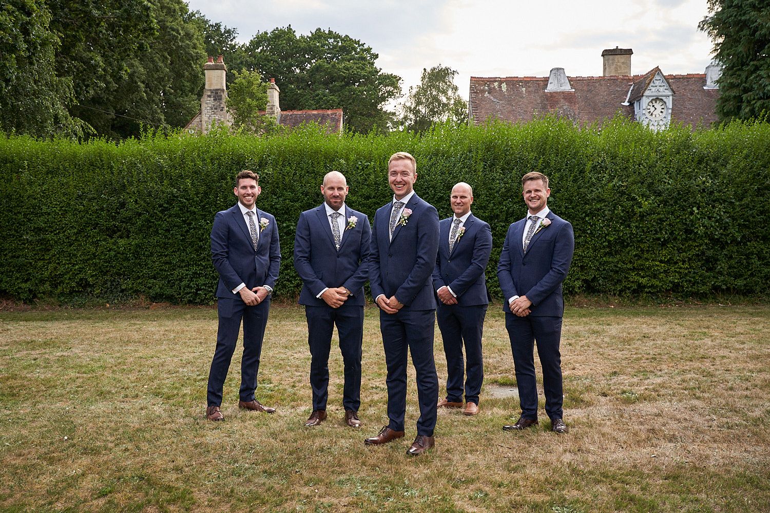 A group of four groomsmen in matching navy blue suits and brown shoes pose in front of a hedgerow at an outdoor venue.