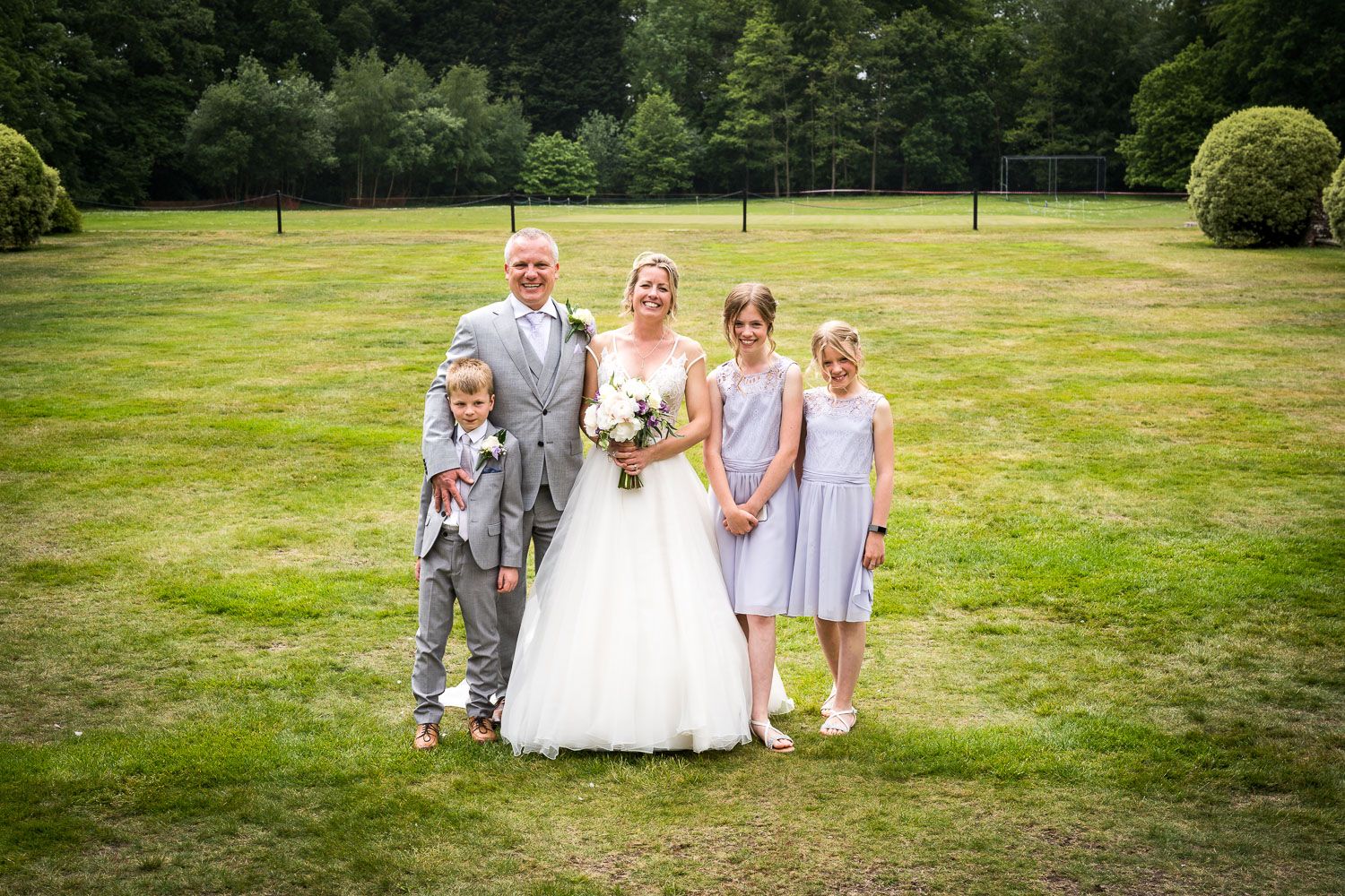 A wedding party poses together on a sunny day in a lush green garden setting.