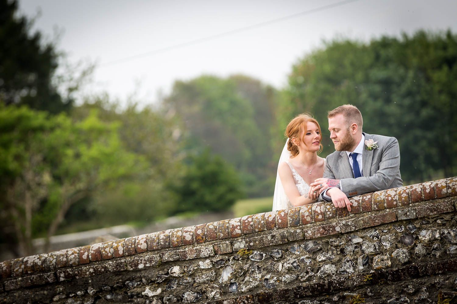 A romantic couple shares a tender moment while leaning on an old stone wall surrounded by lush greenery on their wedding day.