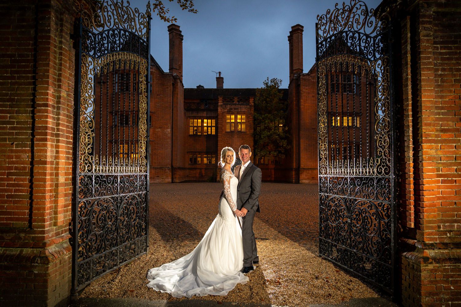 Wedding couple posing at ornate iron gates of illuminated historic brick mansion at night.