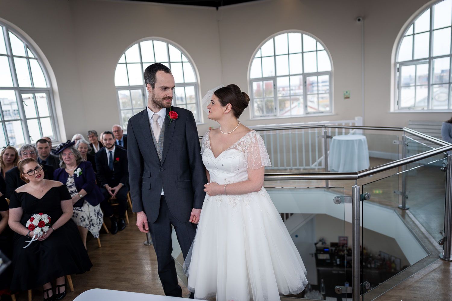 Couple exchanges meaningful glance at wedding ceremony in bright indoor venue with arched windows and guests.