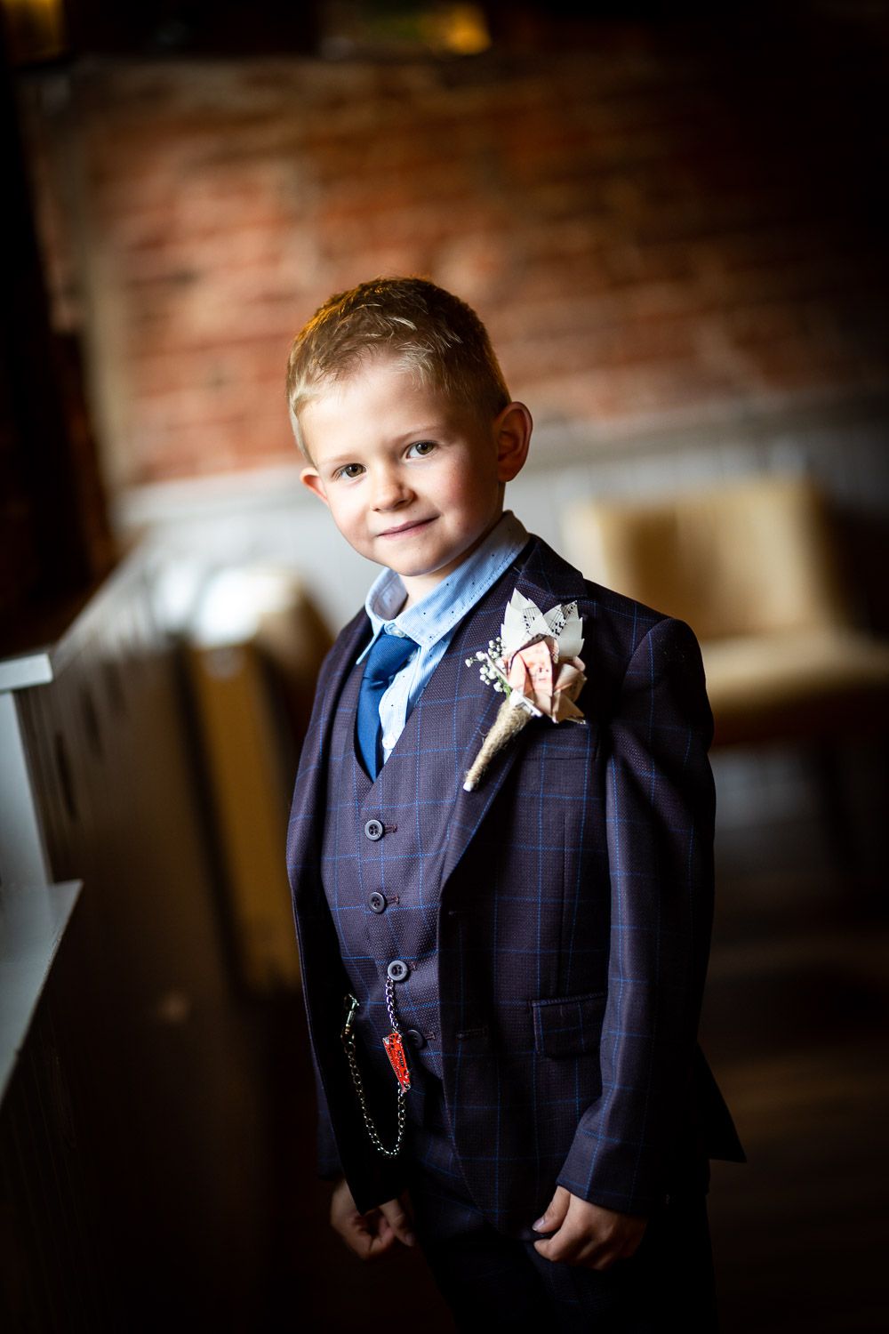 A young attendant at a formal event wearing a navy three-piece suit with boutonniere in front of a brick wall.