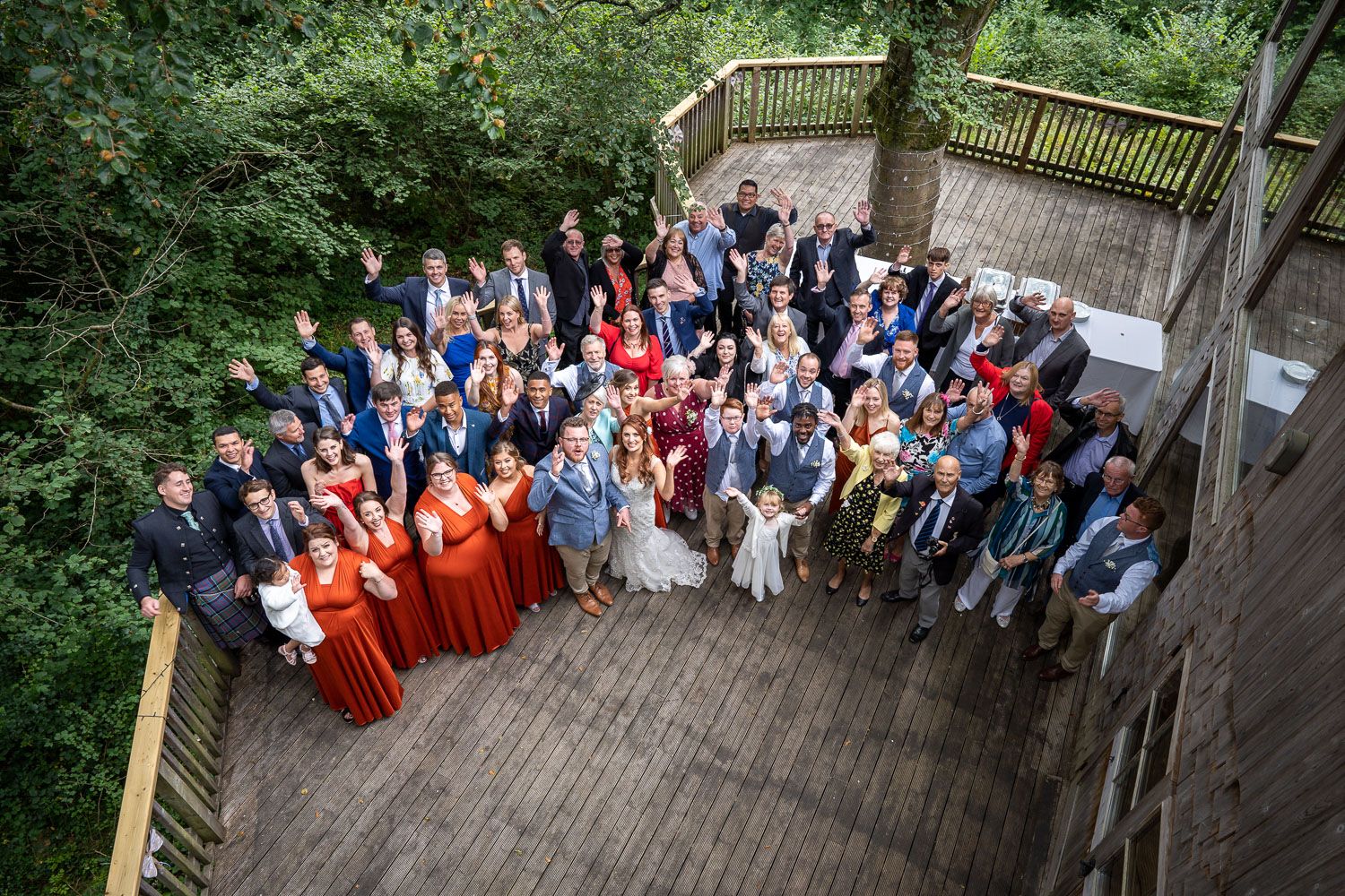 Aerial view of large wedding party gathered on outdoor wooden deck surrounded by lush green forest.