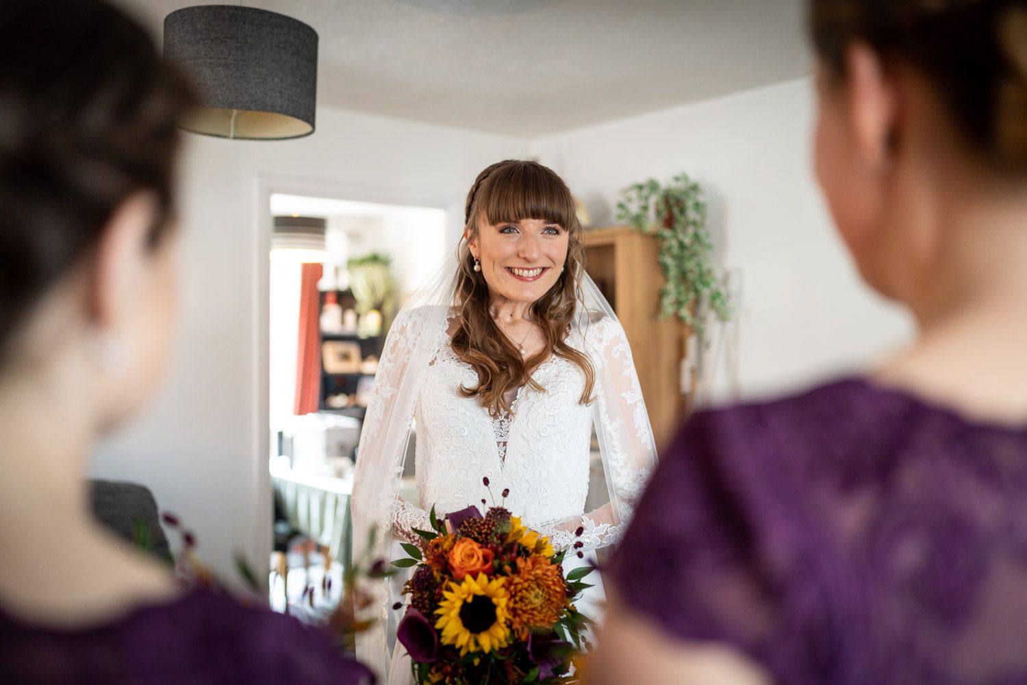 A bride holding a sunflower bouquet smiles during an indoor wedding preparation scene.