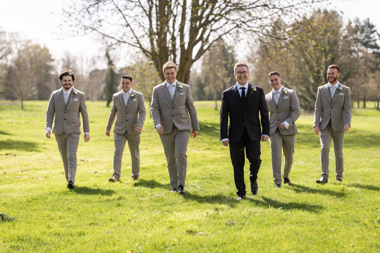 Groomsmen in light gray suits and one in black walk across a sunny green lawn during a wedding celebration.