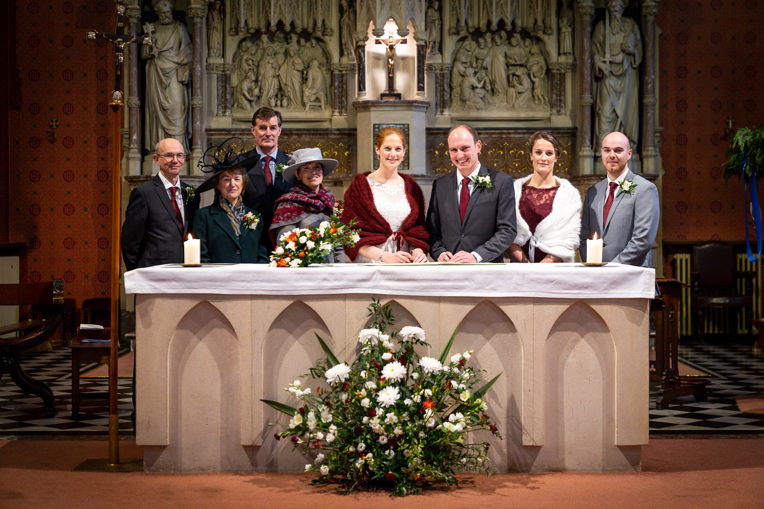 A wedding group stands at the church altar behind a decorative stone railing adorned with floral arrangements.