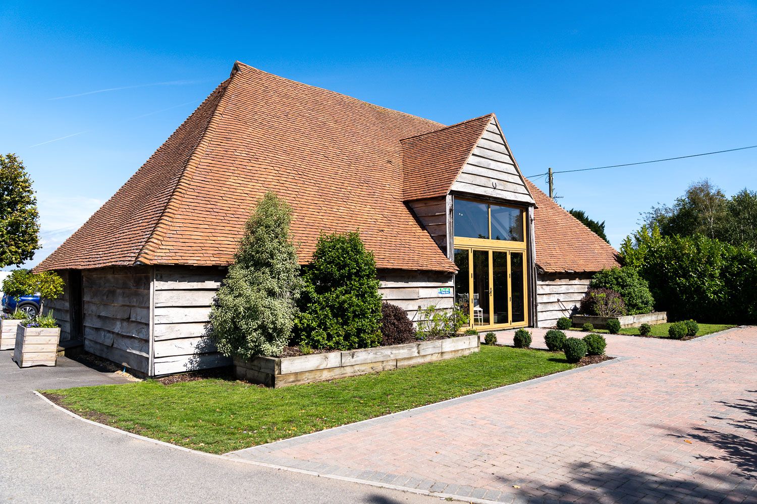 Rustic converted barn with white-painted timber siding, large glass windows and terracotta roof tiles set against blue sky.