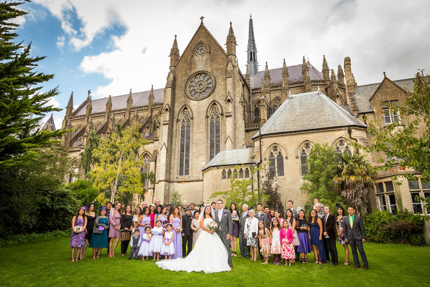 Large group poses for wedding photo outside grand gothic cathedral with ornate spires and beautiful grounds.