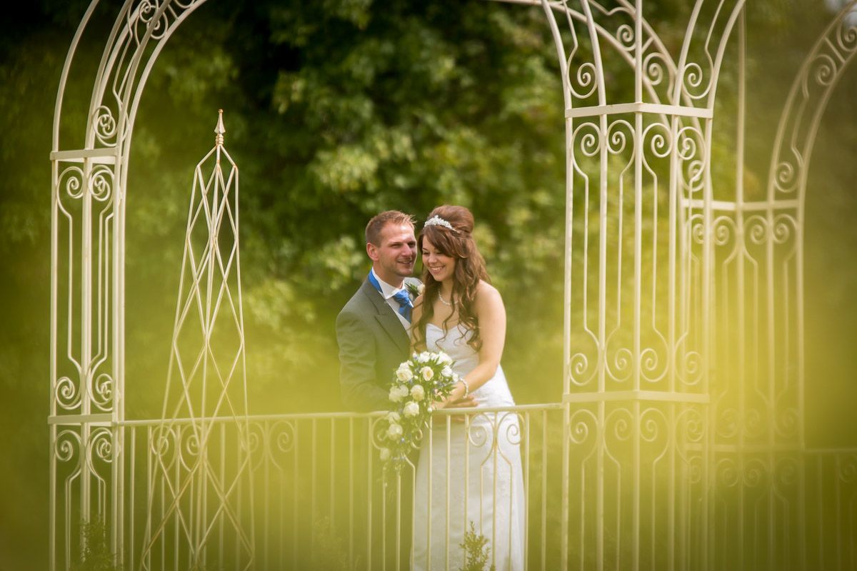 A romantic wedding couple stands beneath an ornate white metal garden arch surrounded by soft green foliage.