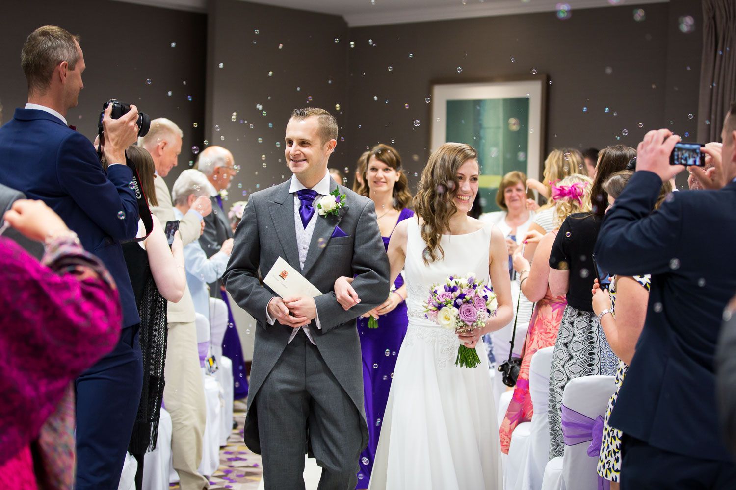 Guests throw confetti over a newlywed couple leaving their wedding ceremony at an indoor venue.