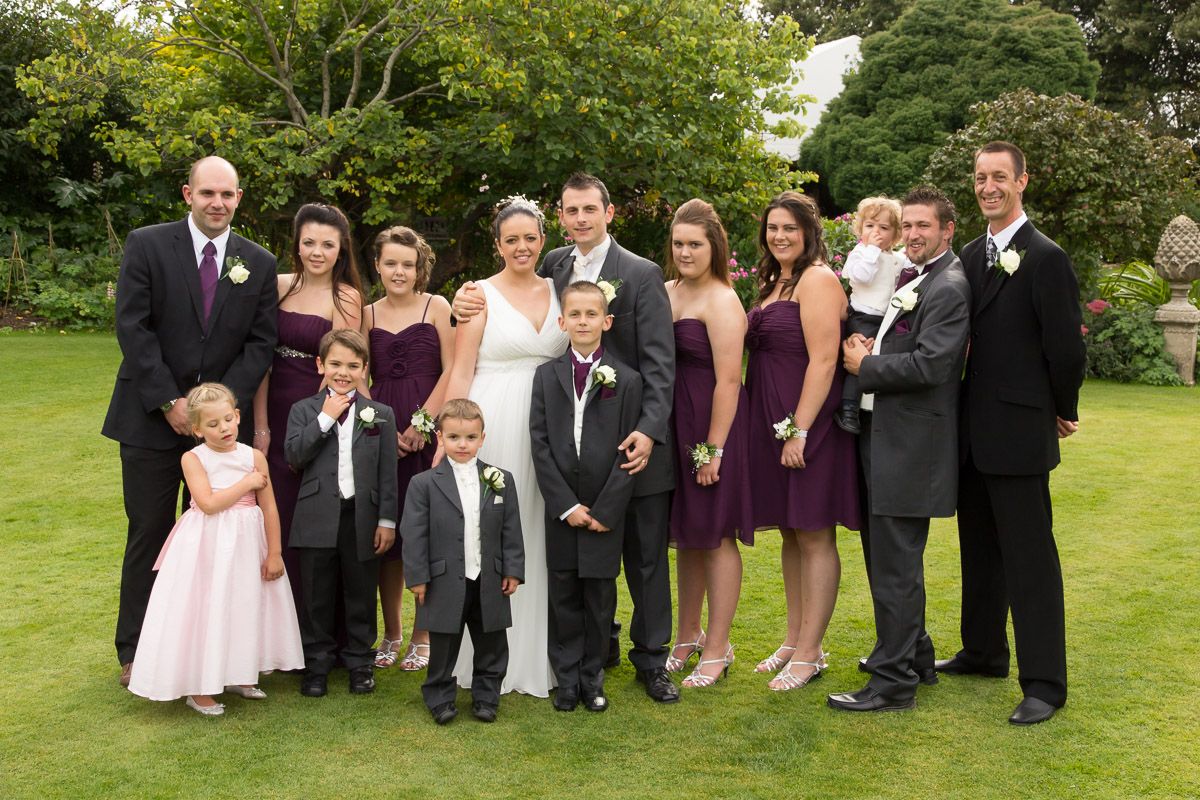 A formal wedding party group poses together on a lawn wearing purple dresses and black tuxedos.