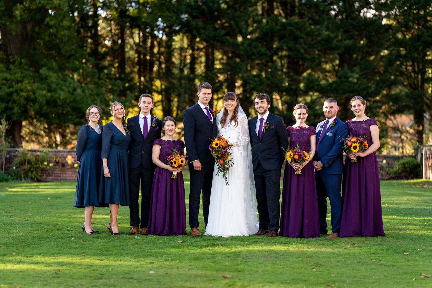 Wedding party in navy suits and purple dresses poses together on green lawn with forest backdrop.