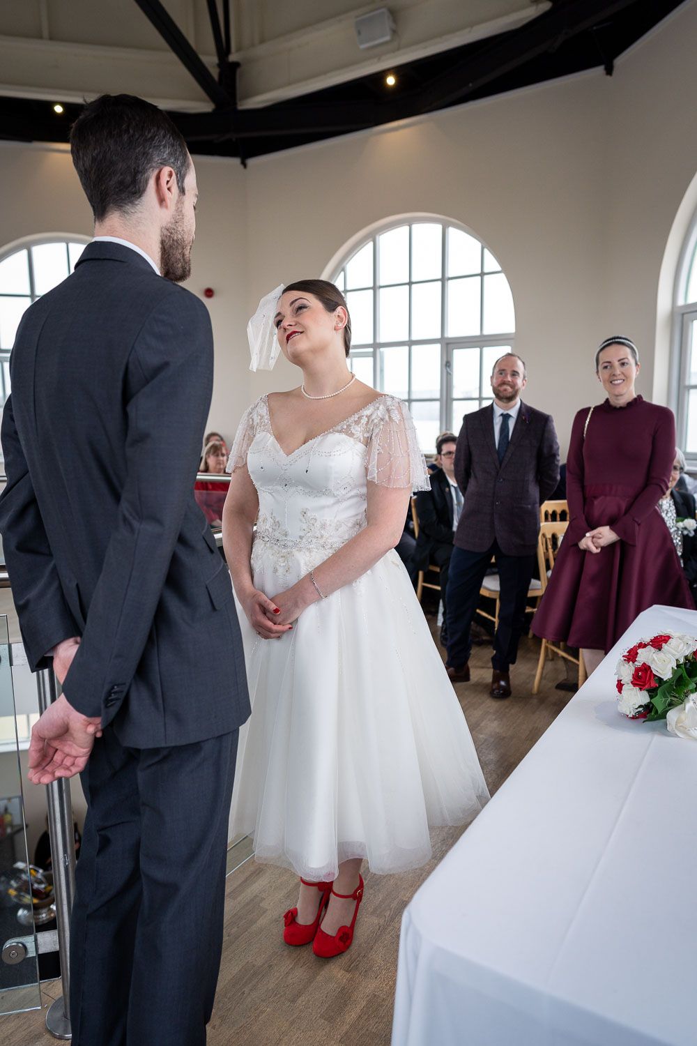 Wedding ceremony in a bright venue with arched windows showing a couple in formal attire with guests looking on.