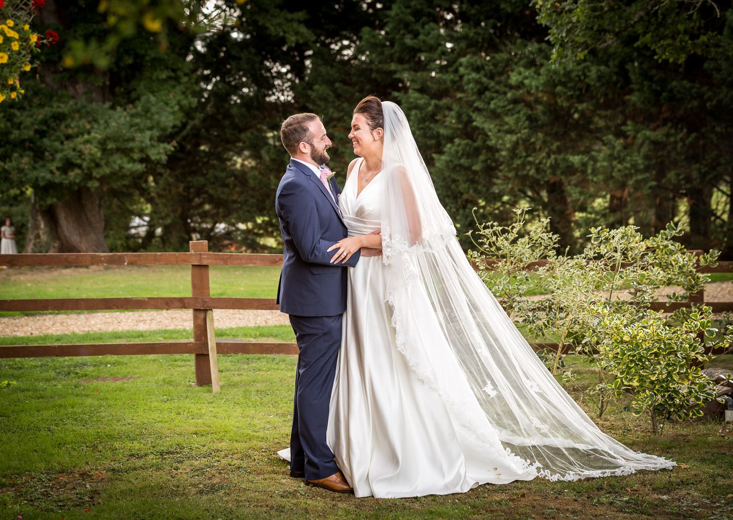 A couple in wedding attire embrace outdoors in a garden setting with a rustic wooden fence and lush greenery.