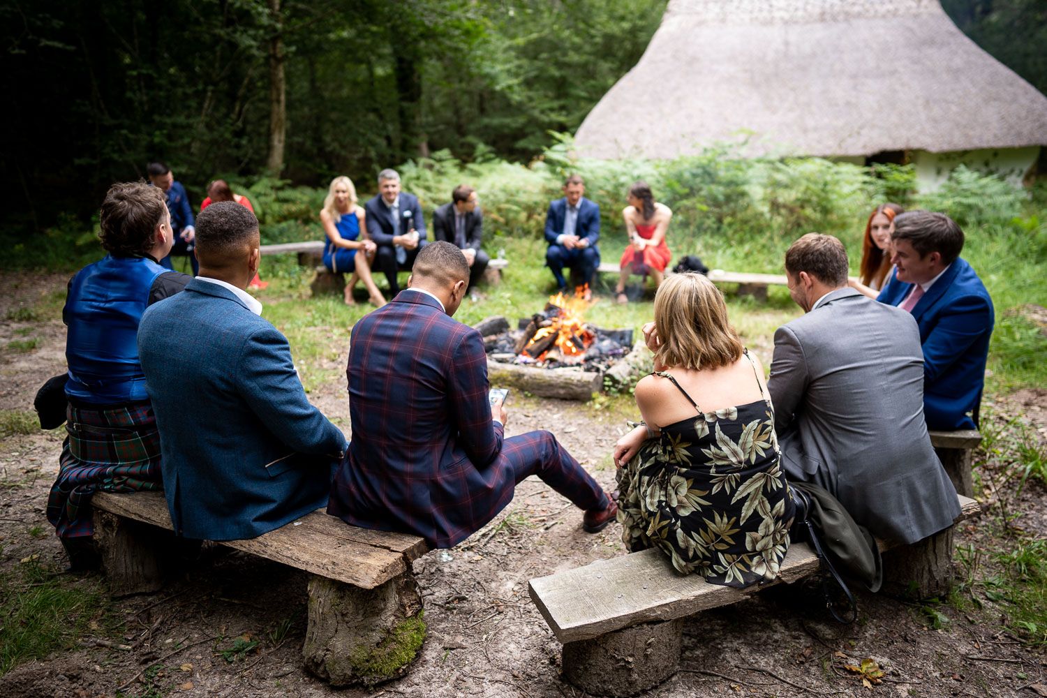 A group of people sit around a campfire outside a thatched-roof hut in a wooded area during a gathering.