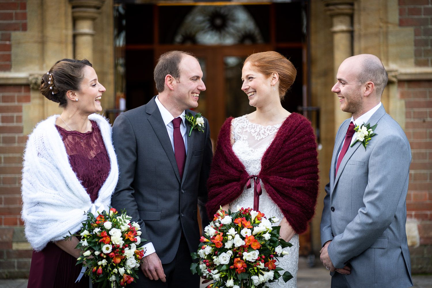 A wedding party in burgundy and white attire poses together outside a brick church entrance.