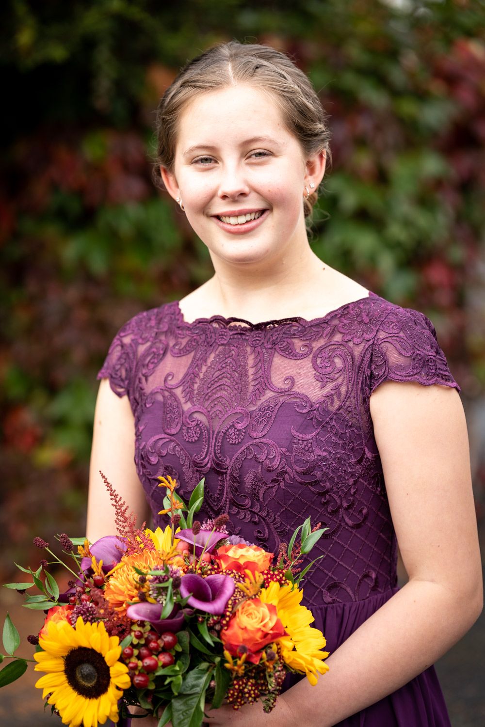Bridesmaid in purple lace dress holds vibrant fall bouquet with sunflowers, roses and berries against autumn foliage.