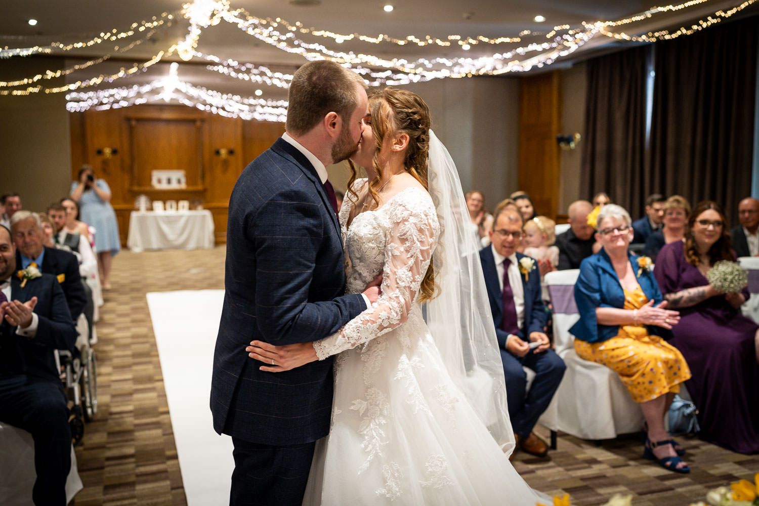 At an indoor wedding reception, a couple shares their first kiss under twinkling string lights as guests look on.