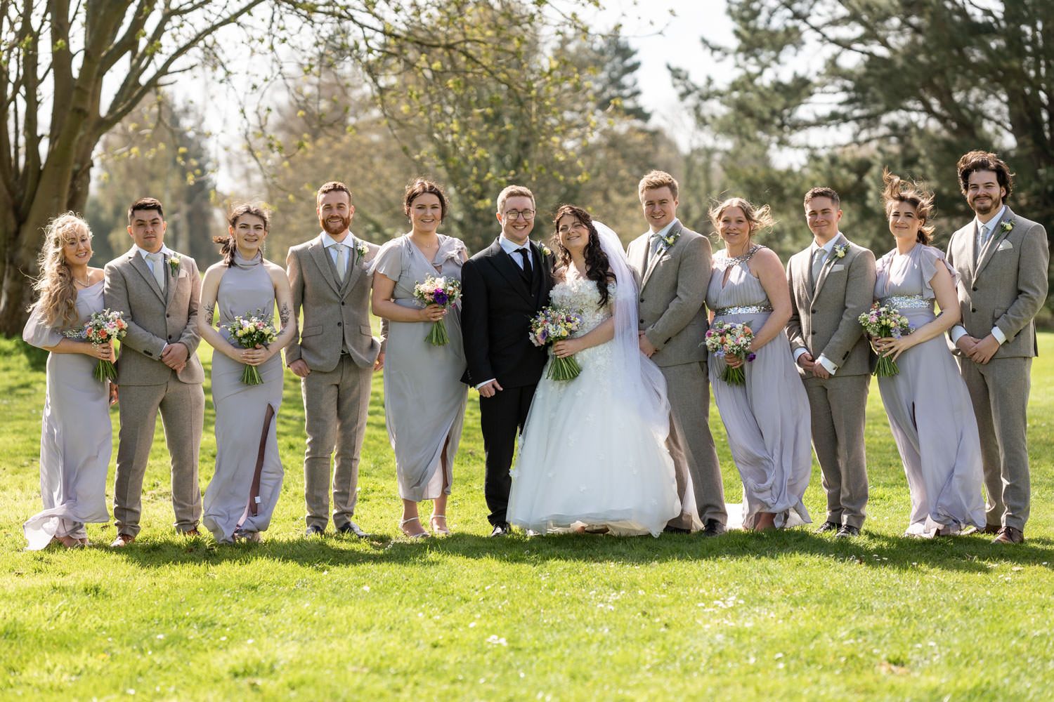 Wedding party in light gray suits and a white gown pose together on sunny grass lawn surrounded by trees.