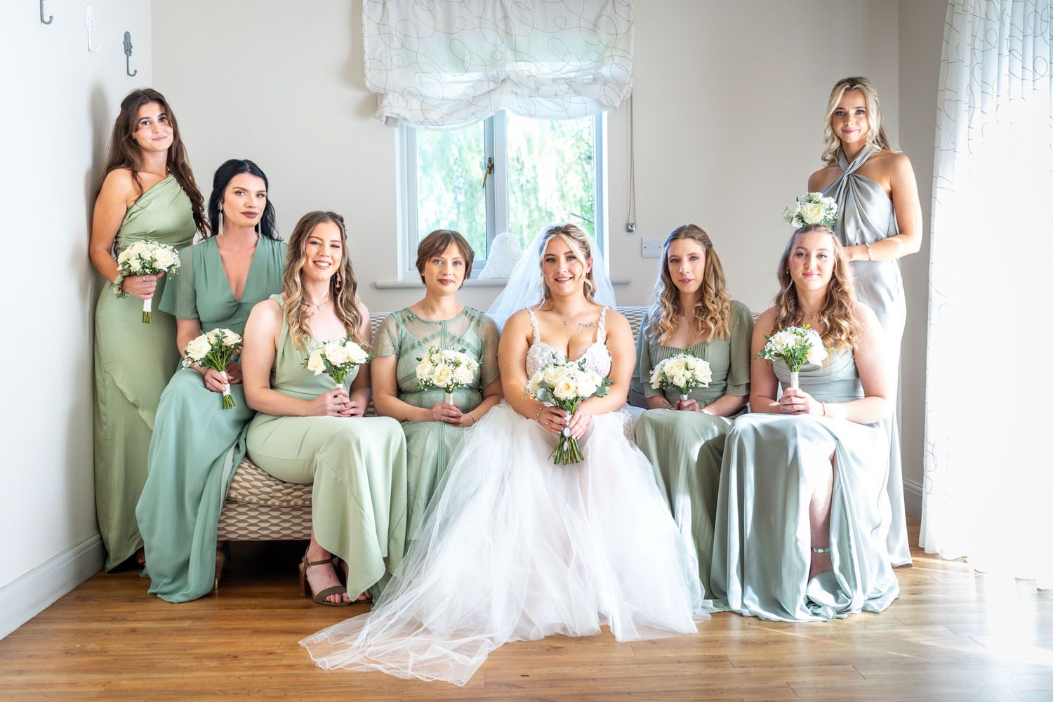 Bride poses with six bridesmaids in sage green dresses, holding white bouquets in a bright white room.