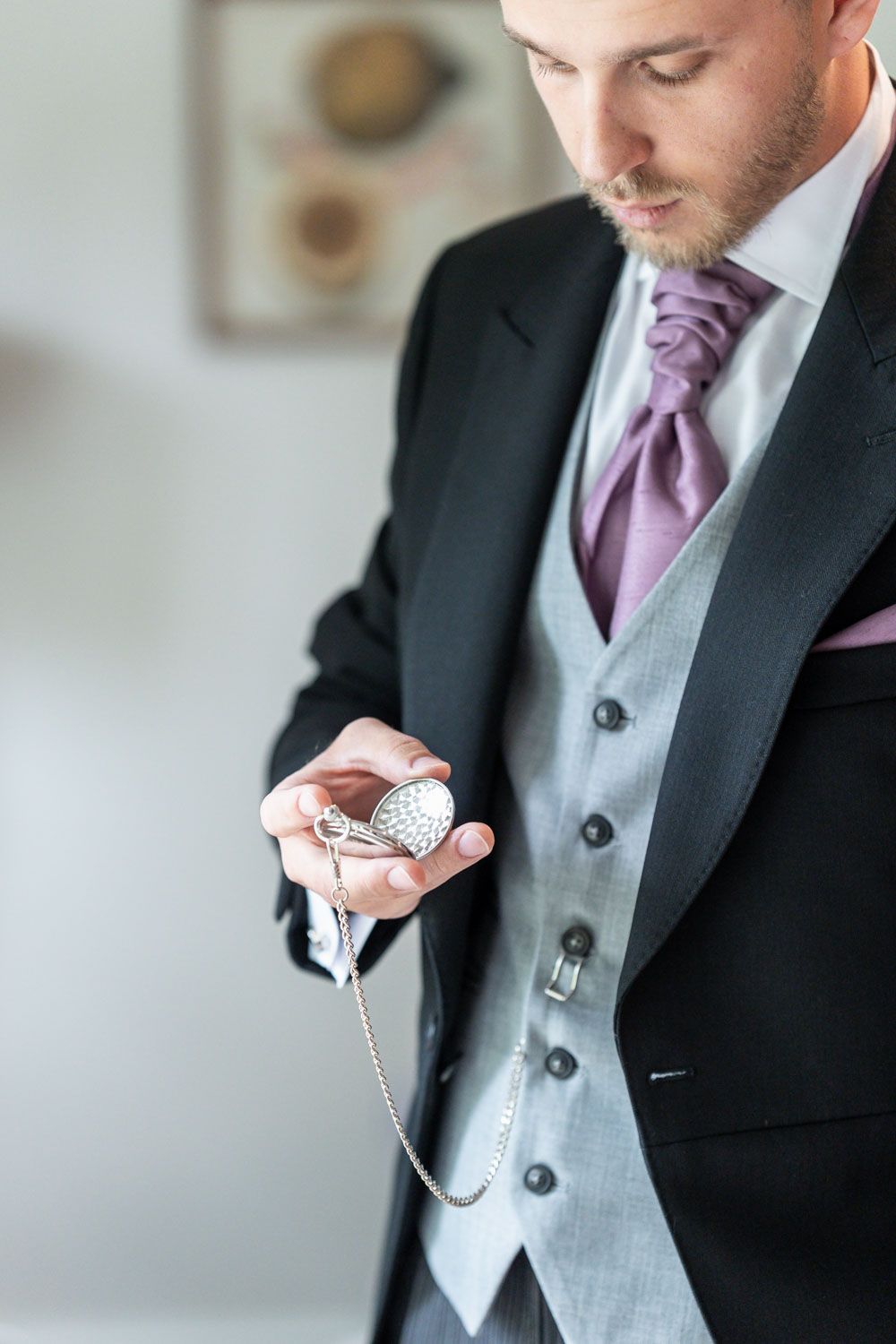A groom in formal attire with a black suit and purple tie holds an antique pocket watch before the ceremony.