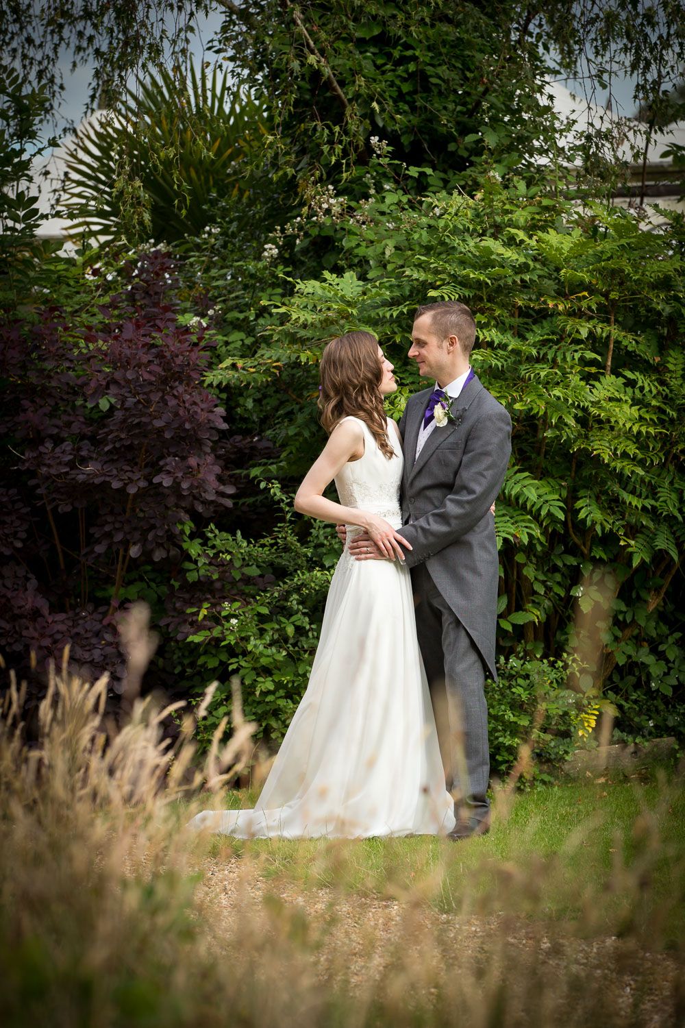 A couple in wedding attire embrace in a lush garden setting with vibrant greenery and wildflowers.