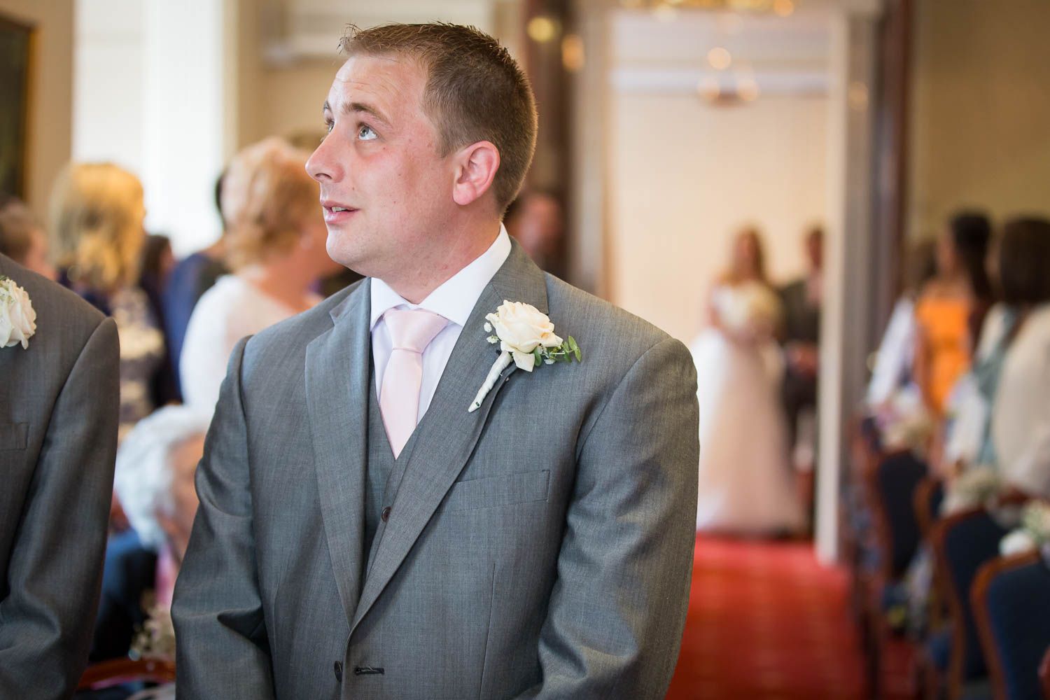 A groom in a light gray three-piece suit with white boutonniere smiles during a wedding ceremony.