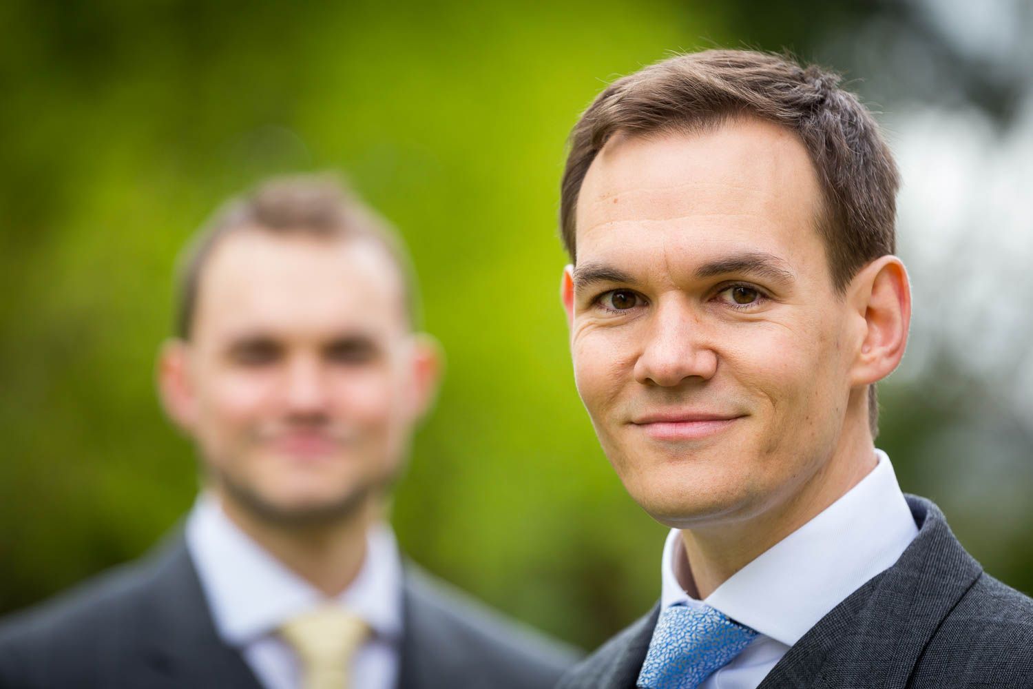 Two men in formal business attire with suits and ties stand outdoors with a blurred green background.