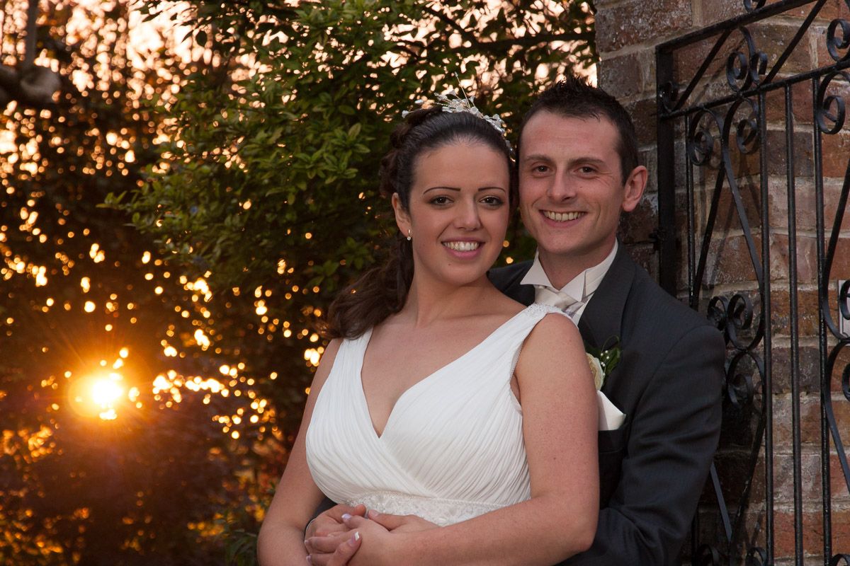 A couple poses for a romantic sunset wedding photo against a stone wall with decorative ironwork.