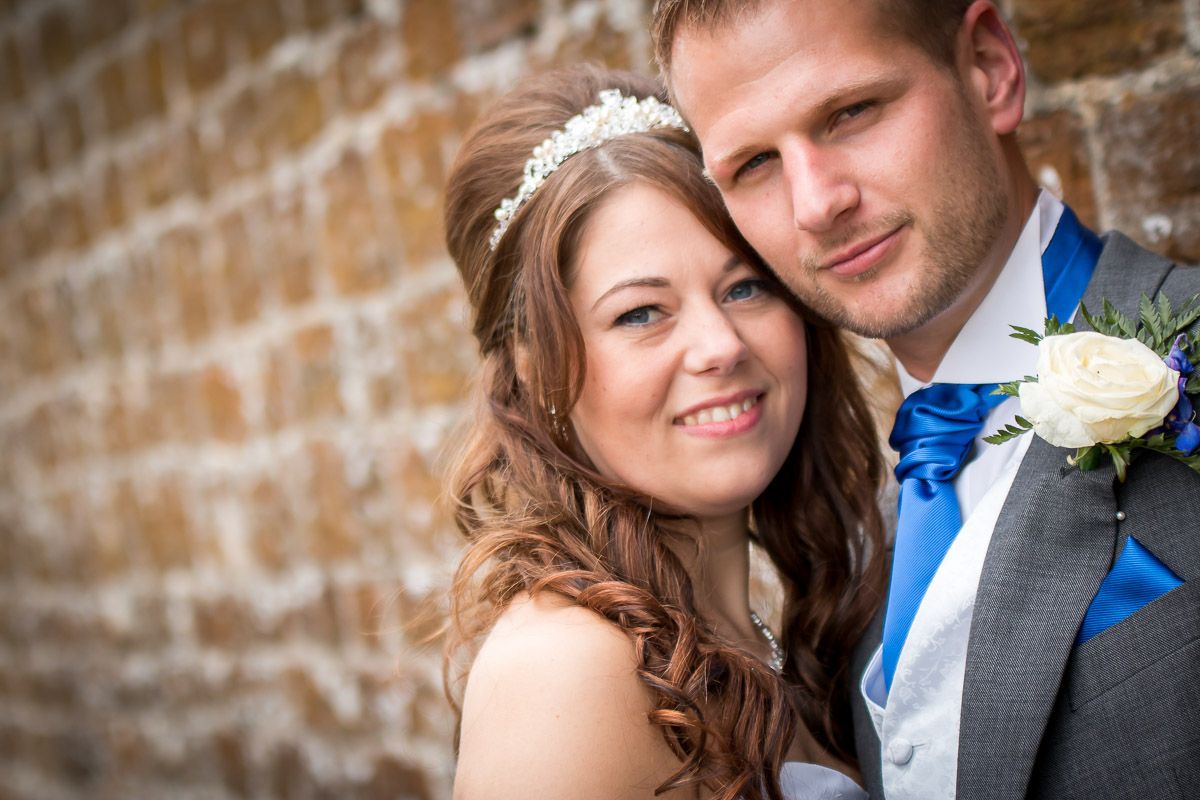 Newlyweds pose together in front of brick wall with groom wearing grey suit with blue tie and bride in white gown.