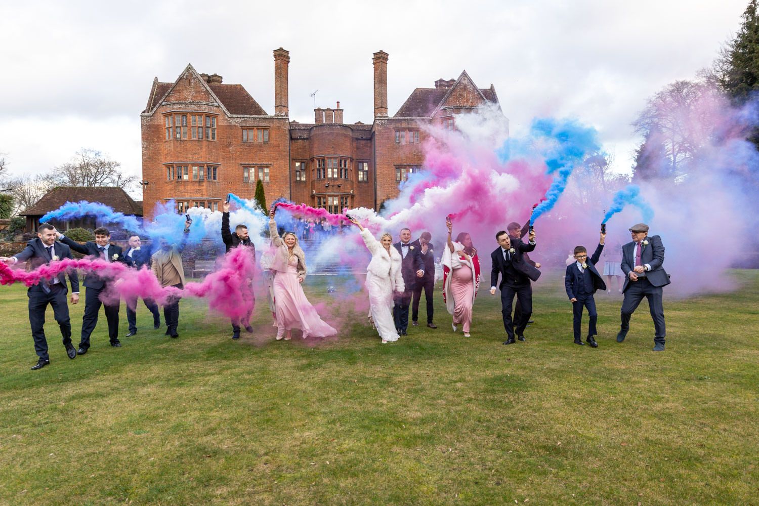Wedding party celebrating with colorful pink and blue smoke bombs on the lawn of a grand manor.