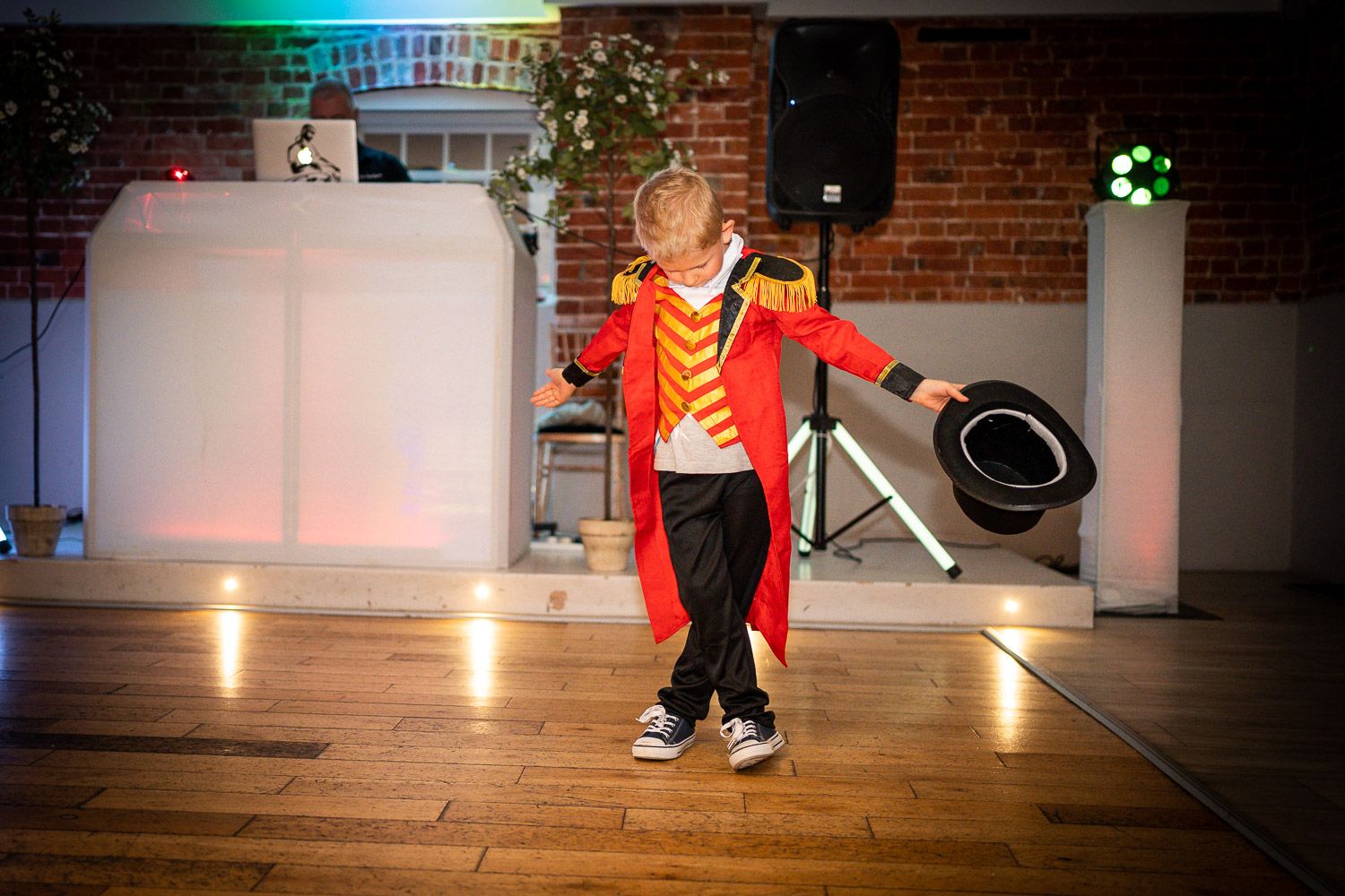 A young dancer performs in a bright red and gold costume on a wooden dance floor at an indoor venue.