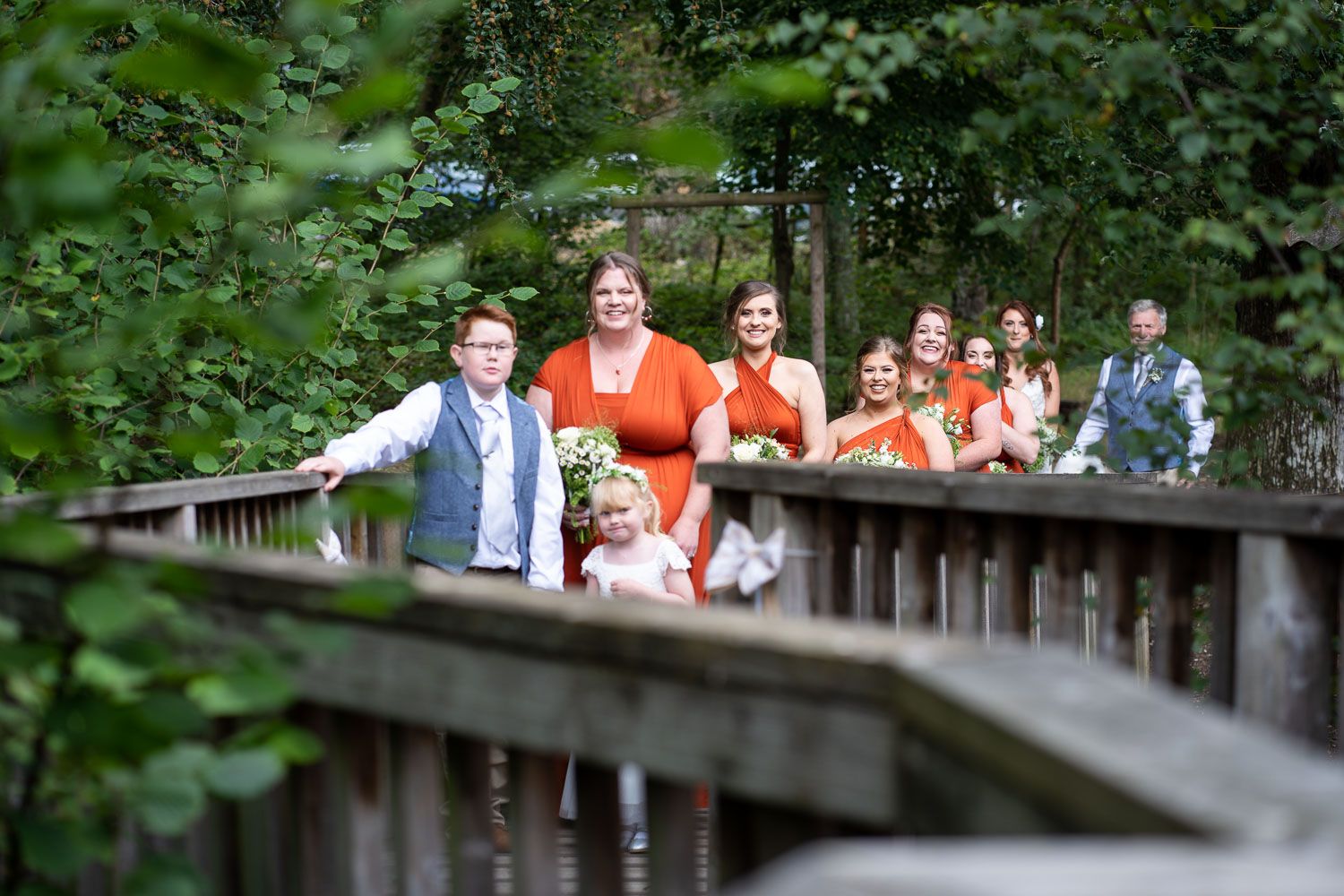 A group of people in colorful attire stand together on a wooden boardwalk path surrounded by green foliage.