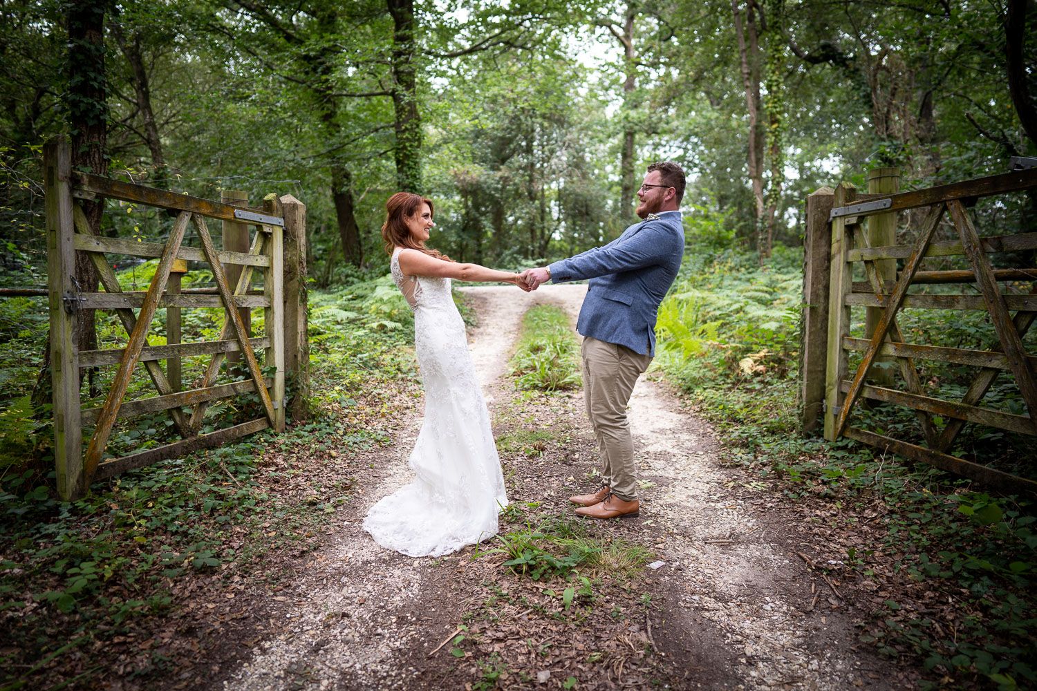 A couple dances together on a dirt path between rustic wooden gates in a lush green forest setting.