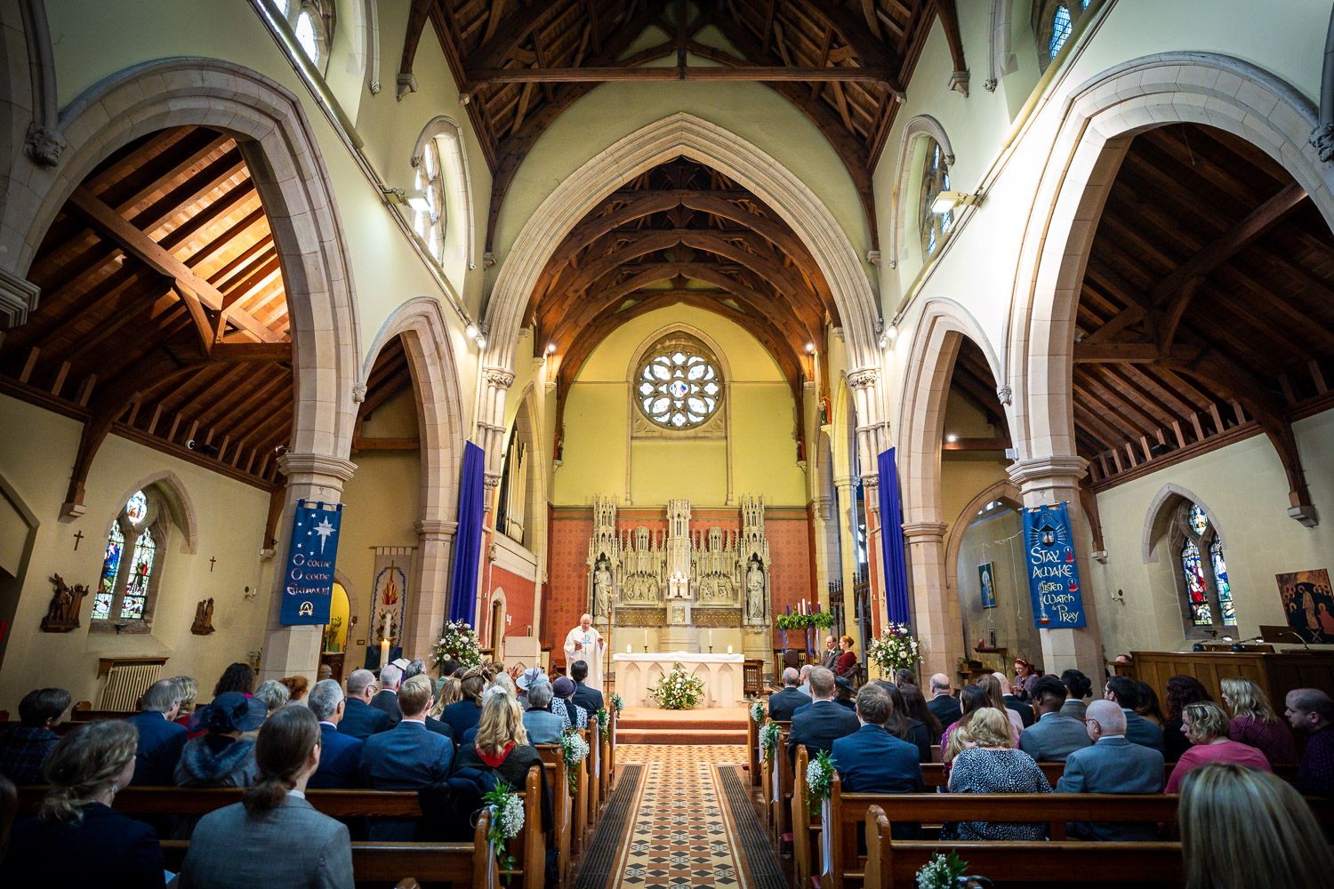 Interior view of a church during a service with high vaulted arches, stained glass windows and wooden beams.