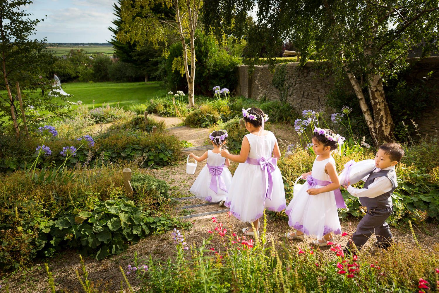 Small children in white dresses walking through a lush garden with blooming flowers and greenery.
