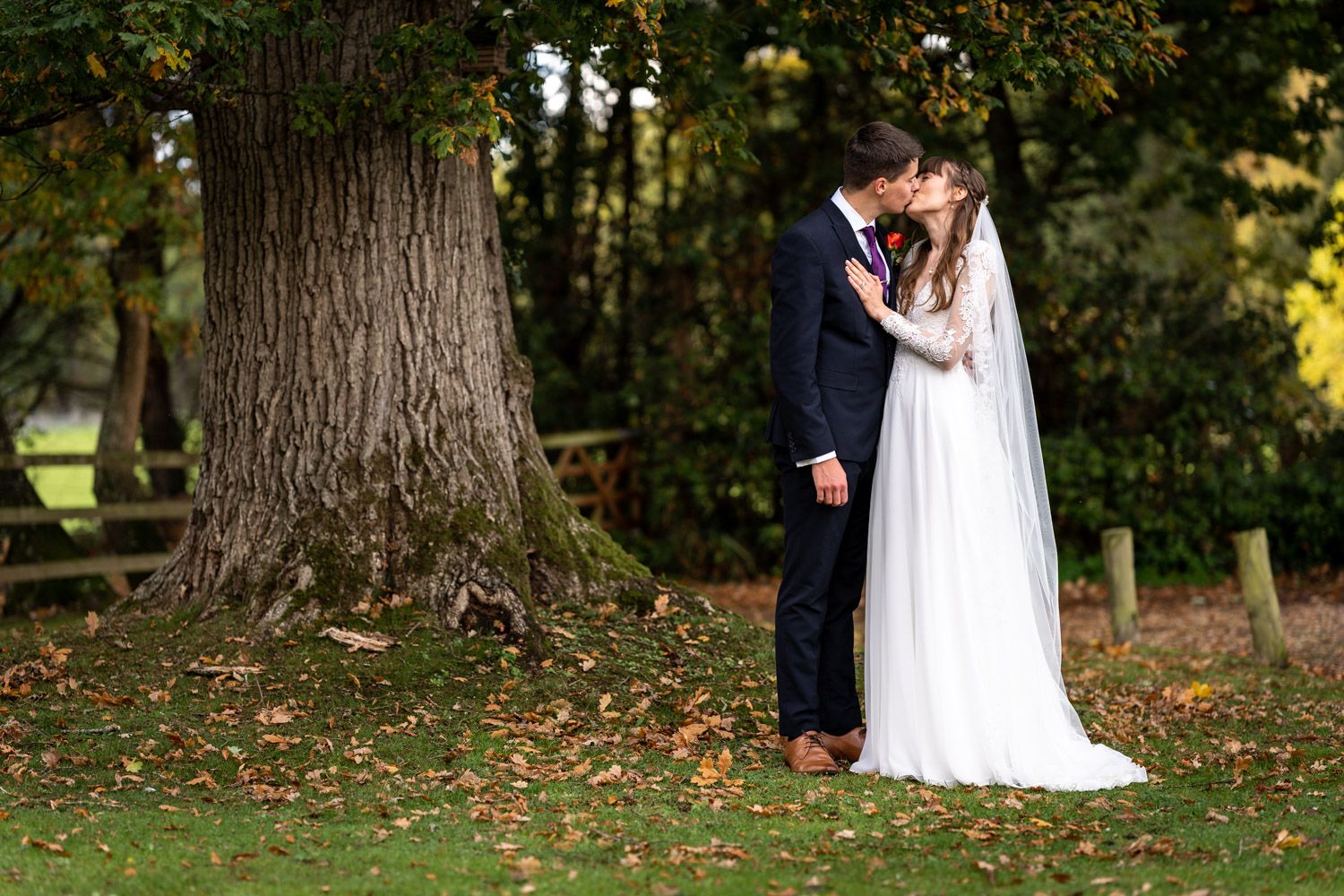 A romantic wedding couple shares an intimate moment on fall grass near a large tree trunk in an outdoor setting.